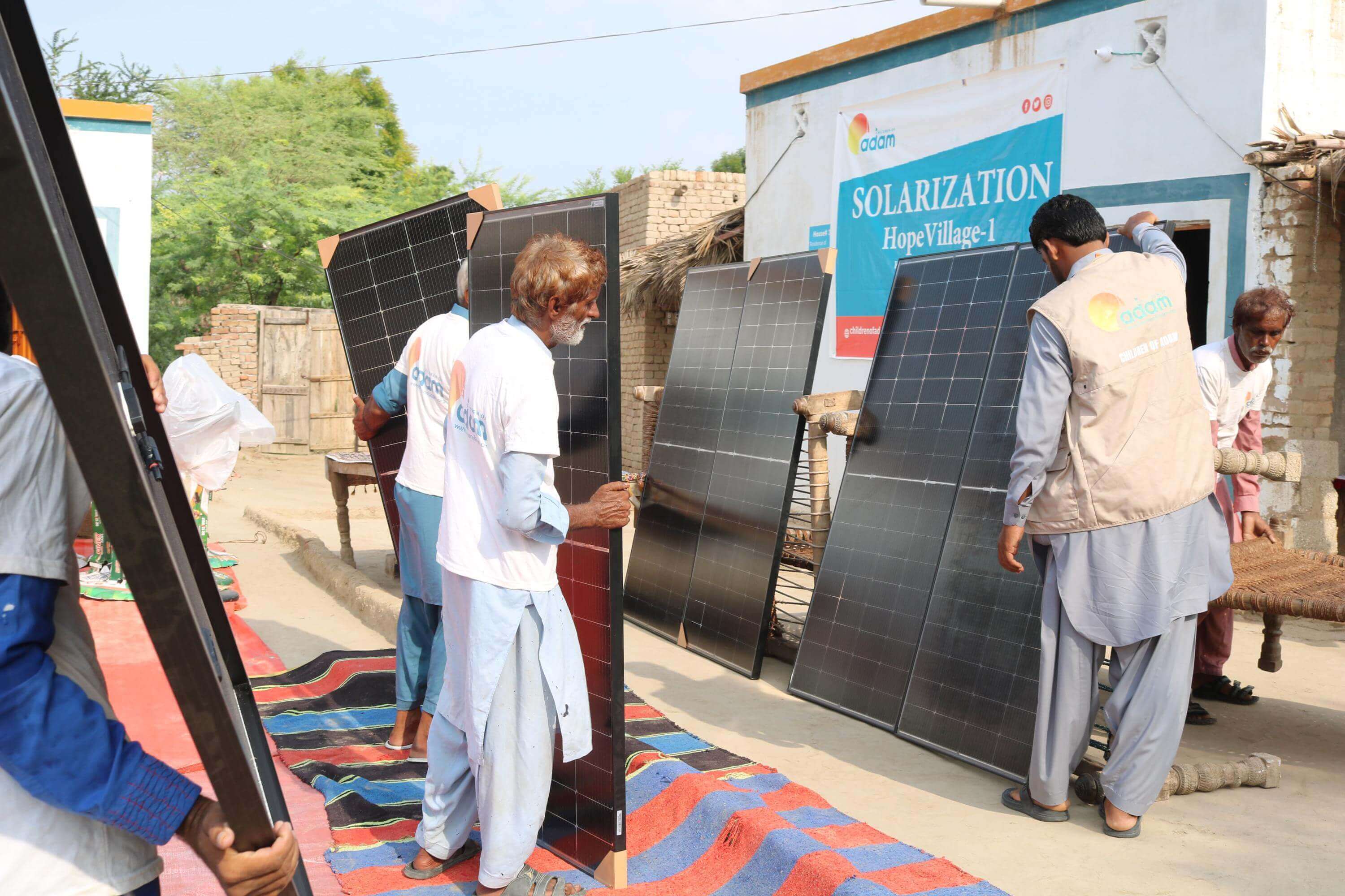 Pakistan Heatwave Appeal: How You Can Help Families Survive the Summer 1 A group of men are working together to set up several large solar panels outdoors in a village setting, with a sign in the background reading "solarization hopevillage-1."