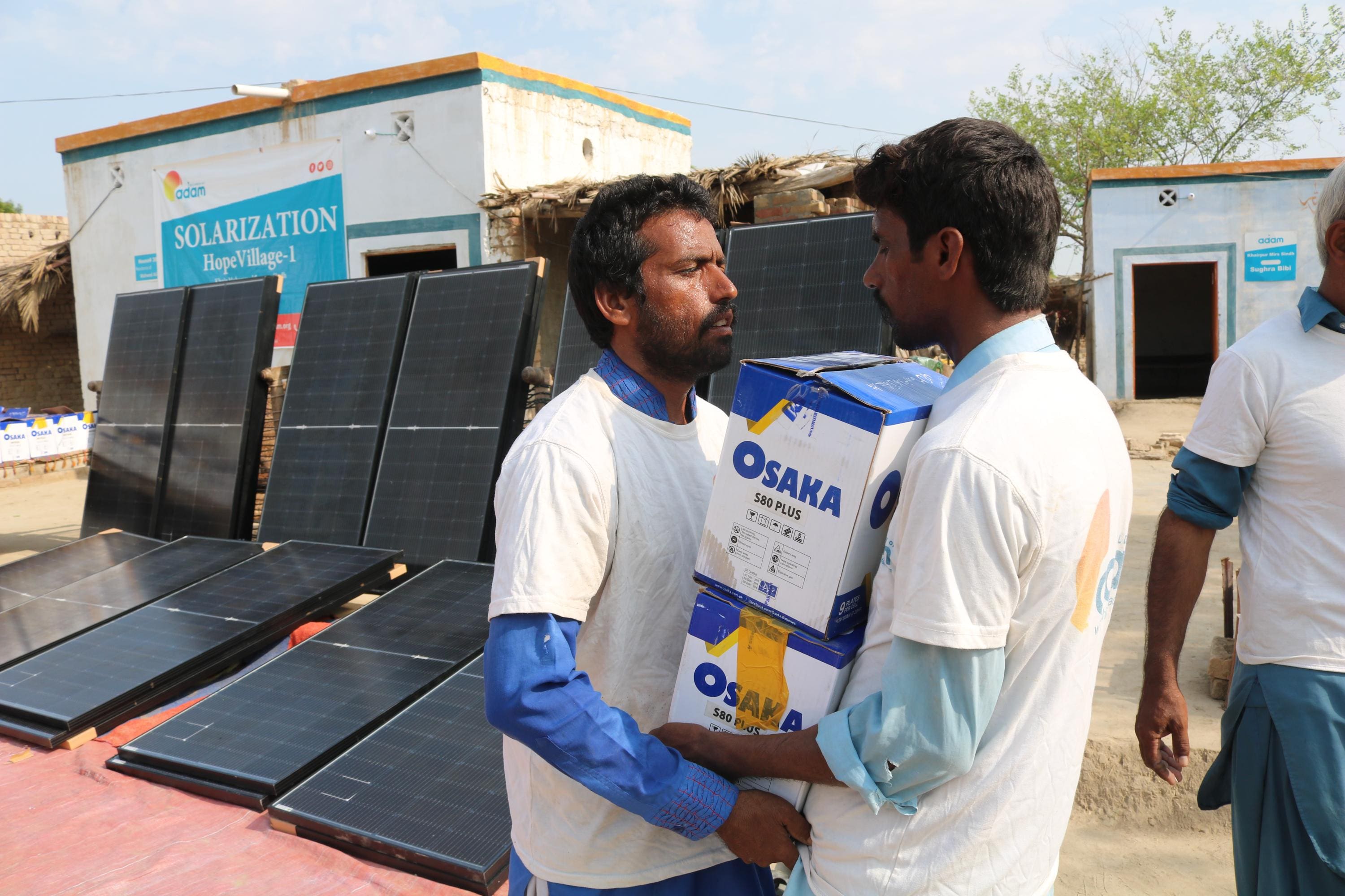 Pakistan Heatwave Appeal: How You Can Help Families Survive the Summer 2 two men carry boxes labeled "osaka s80 plus" in front of several large solar panels laid out in a village setting.