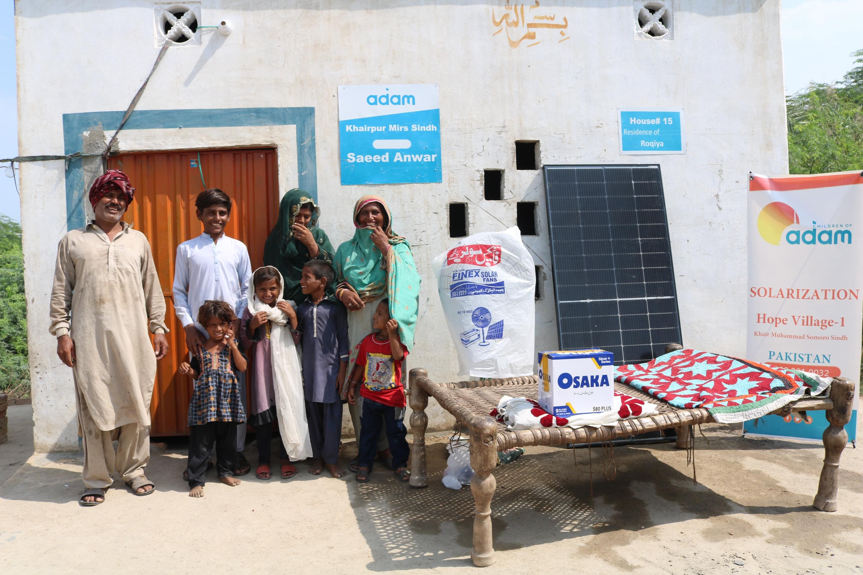 Donate Solar Fans for Families in Pakistan 2 a smiling family poses outside their home in khairpur mirs, sindh, next to a solar panel, an osaka battery box, and a standing fan.