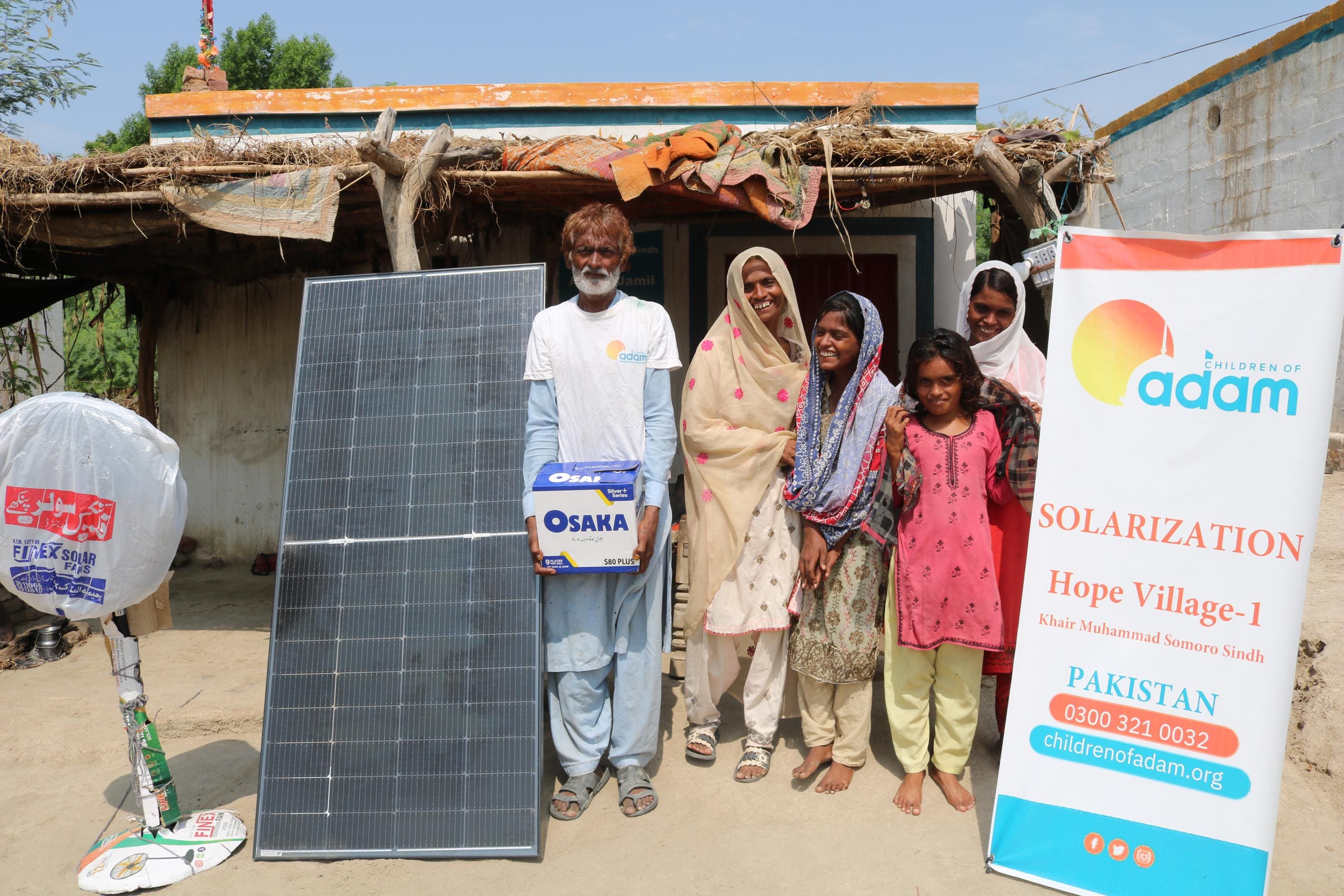 How Power Cuts Make Heatwaves More Dangerous in Pakistan 1 a smiling family poses outside their home in khairpur muhammad somoro, sindh, next to a solar panel, an osaka battery box, and a standing fan.