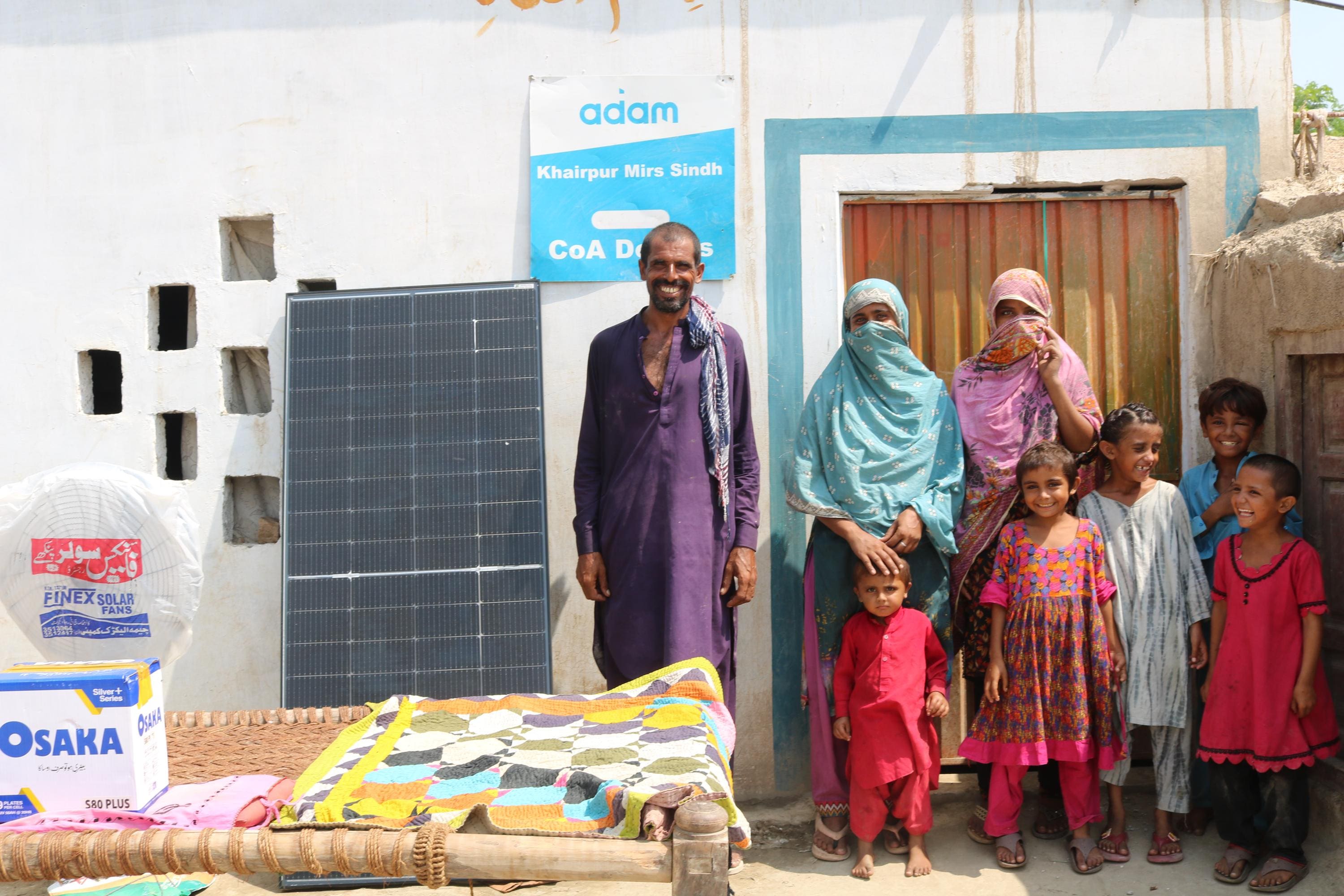 Why Solar Panels Are Sadaqah Jariyah 1 A smiling family poses outside their home in Khairpur Mirs, Sindh, next to a solar panel, an Osaka battery box, and a standing fan.