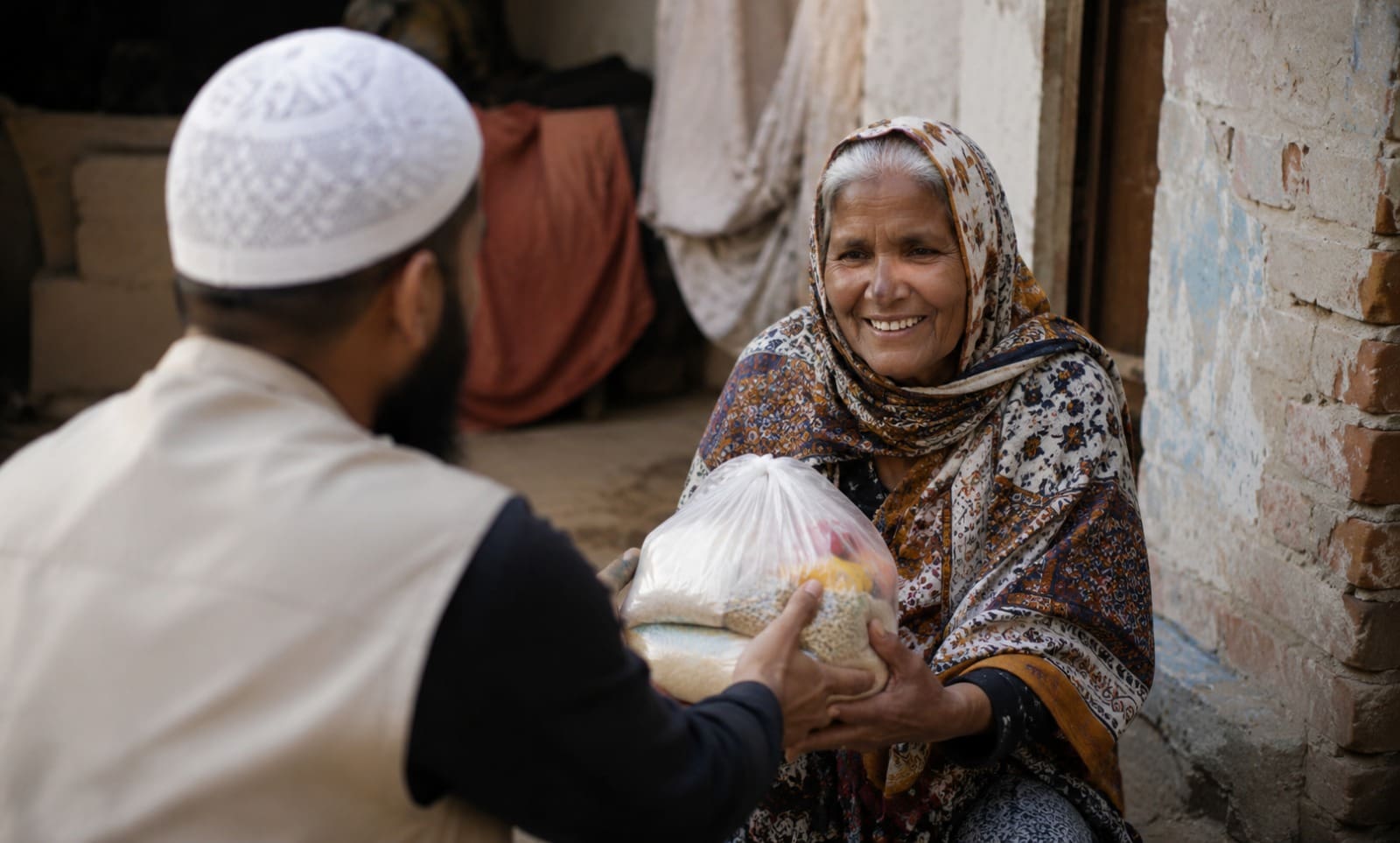 Why Choose a UK Zakat Charity 5 A volunteer handing a bag of food aid to a smiling elderly woman wearing a patterned headscarf