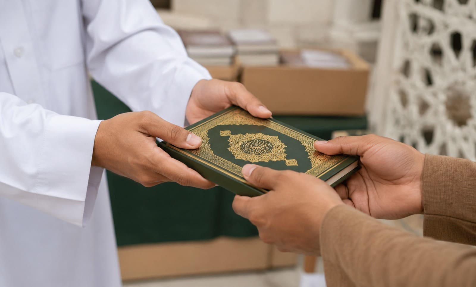 How to Donate a Quran: Step-by-Step Guide 3 a close-up of a person in a white robe passing a green quran with gold arabic calligraphy decoration to another person's hands, with boxes of books blurred in the background.