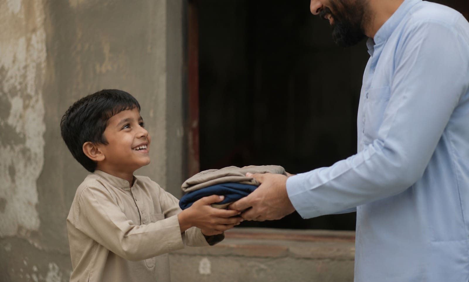 How to Sponsor an Orphan: Everything You Need to Know About This Life-Changing Gift 2 A smiling young boy receiving a stack of folded clothes from a man wearing a light blue kurta.