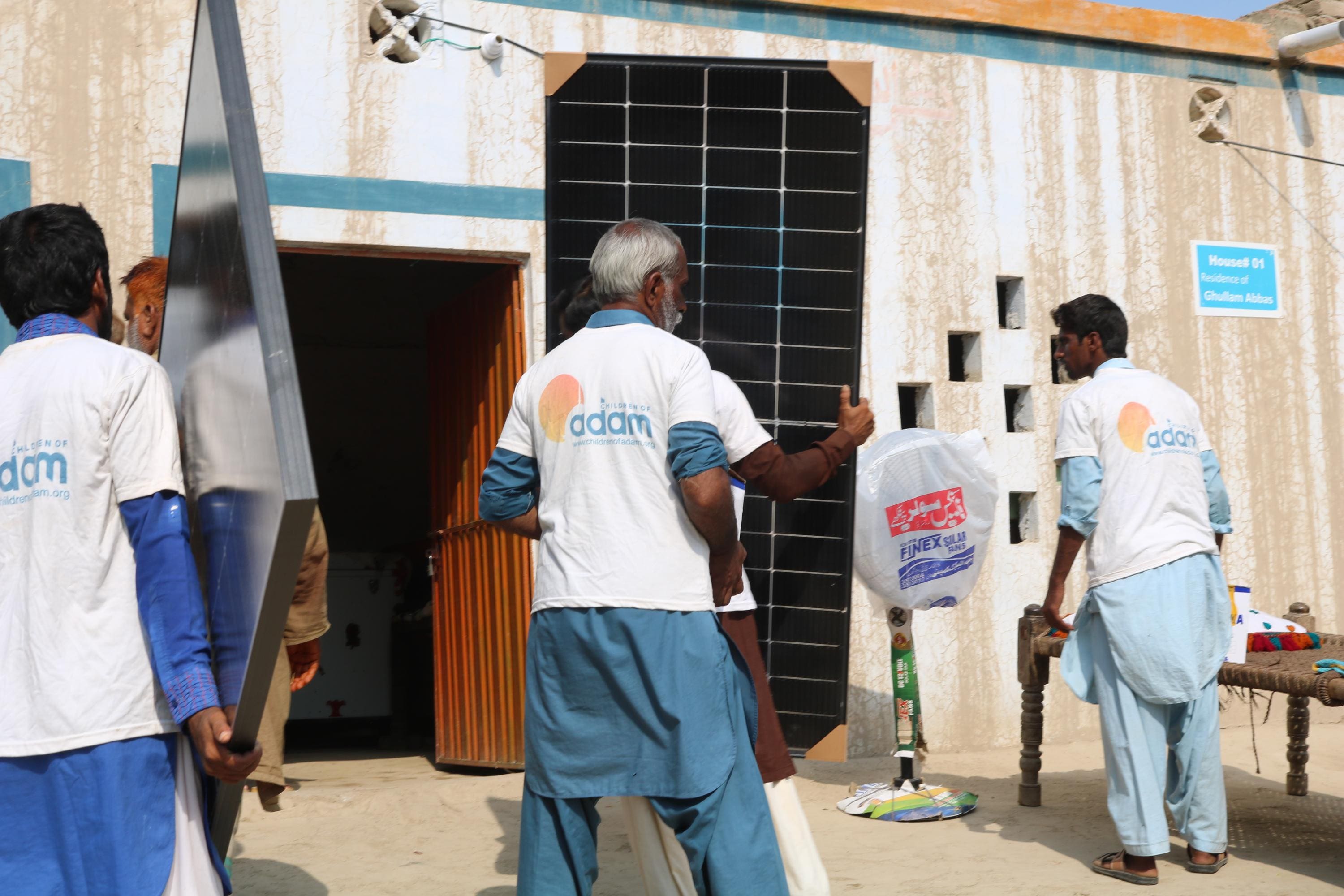 Why Solar Panels Are Sadaqah Jariyah 6 men wearing "children of adam" t-shirts carry and set up solar panels outside a house labeled "residence of ghullam abbas."
